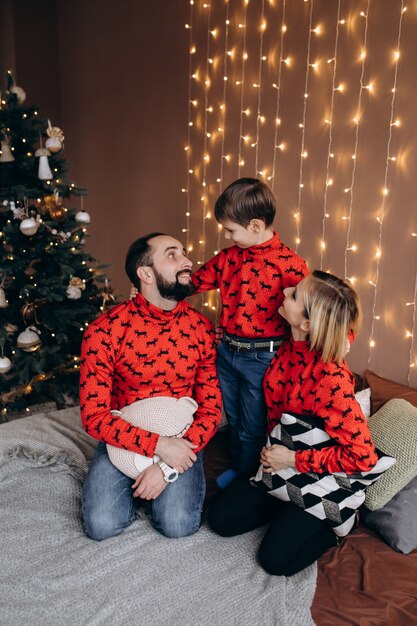 Attractive parents and their little son in red sweaters have fun resting on the bed before a Christm