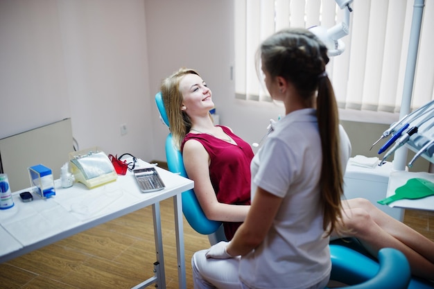 Attractive patient in redviolet dress laying on the dental chair while female dentist treating her teeth with special instruments