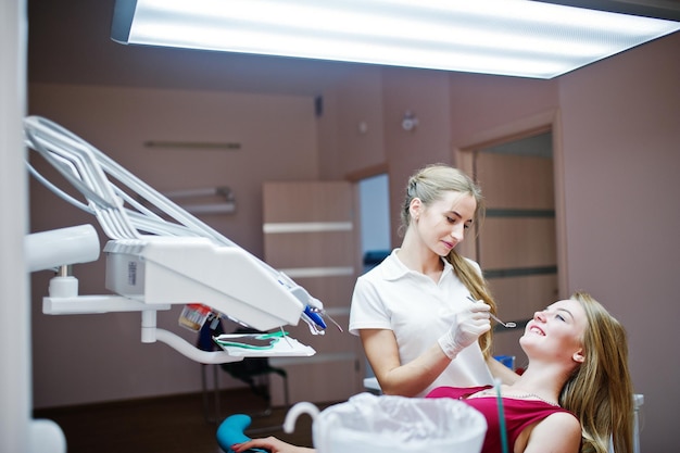 女性の歯科医が特別な器具で彼女の歯を治療している間、歯科用椅子に横たわっている赤紫色のドレスを着た魅力的な患者