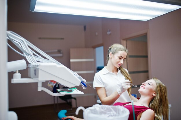 붉은 보라색 드레스를 입은 매력적인 환자가 치과 의자에 누워 있는 동안 여성 치과의사가 특별한 도구로 치아를 치료하는 동안