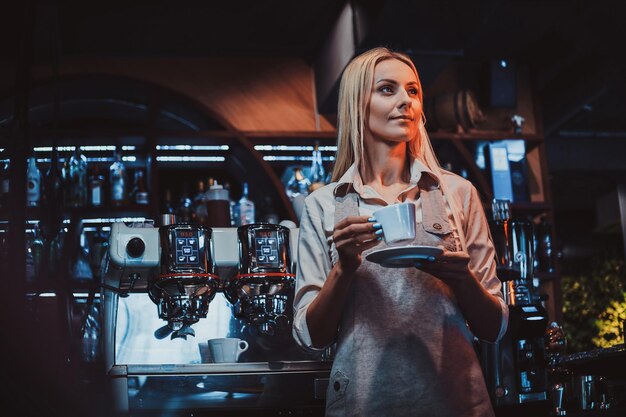 Attractive pensive barista is enjoying her coffee after long shift standing next to coffee maker.