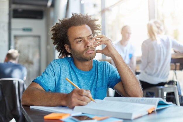 Привлекательный задумчивый молодой студент-мужчина в синей футболке мечтает во время работы над домашним заданием в коворкинг-кафе, пишет сочинение, готовится к английскому или литературе, с задумчивым взглядом