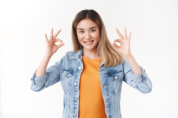 Привлекательная довольная азиатская студентка показывает хорошо, хорошо, жест подтверждения наслаждайтесь идеальной вечеринкой, широко улыбаясь, в джинсовой куртке, оранжевой футболке