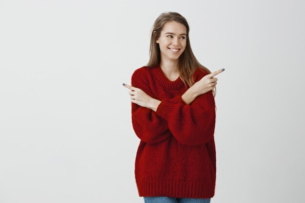 Attractive popular girl making decision on what date go. Pleased joyful caucasian woman in red loose trendy sweater, looking right and pointing in different directions, hesitating with broad smile