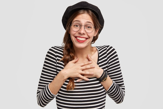 Attractive positive woman dressed in French style, keeps both palms on heart