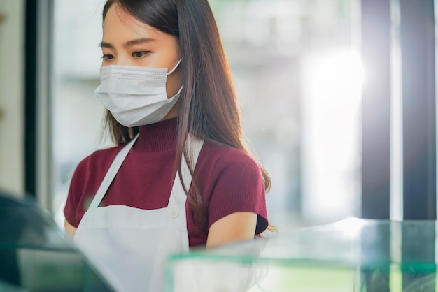 制服のエプロンを着た魅力的なポジティブな若いアジア人女性スタッフが、朝のカフェを安全かつ清潔に消毒するときに、カウンター製品のキャビネットに洗剤またはアルコールをスプレーして柔らかい布で拭きます