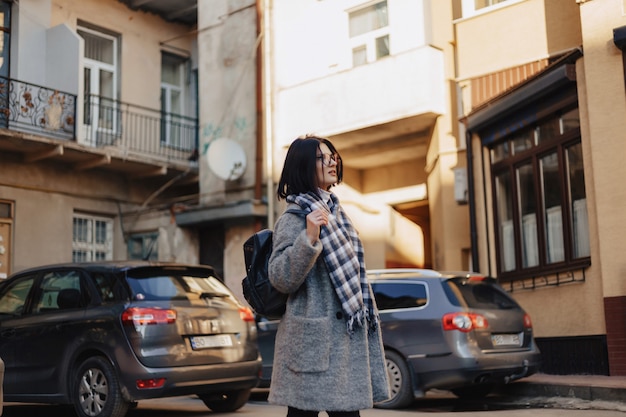 Attractive positive young girl wearing glasses in a coat on the surface of buildings on cars