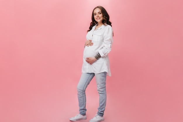 Привлекательная беременная женщина в белой длинной рубашке широко улыбается. Счастливая брюнетка девушка в джинсах позирует на розовом фоне.