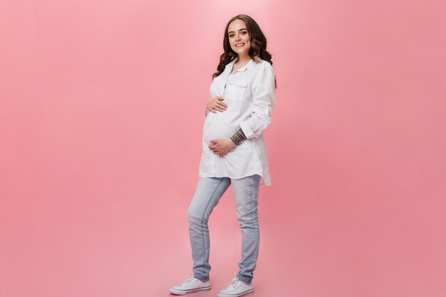 Attractive pregnant woman in white long shirt smiles widely. Happy brunette girl in jeans poses on pink background.