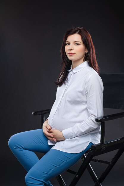 Attractive pregnant woman with dark hair sitting on a black chair and smiling to the camera, picture isolated on black background