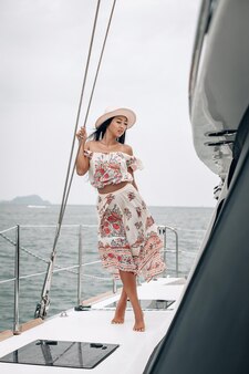 Привлекательная красотка в пляжном платье и панаме стоит на своей частной яхте