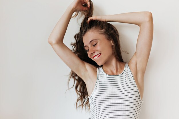 Foto gratuita attraente bella donna con lunghi capelli ondulati che indossa la maglietta spogliata mordendosi un labbro e toccando i capelli sul muro isolato