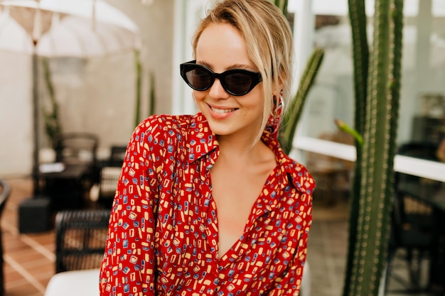 Attractive pretty young woman with blond hair wearing black sunglasses and bright summer dress posing at camera with happy smile on background of summer cafeteria