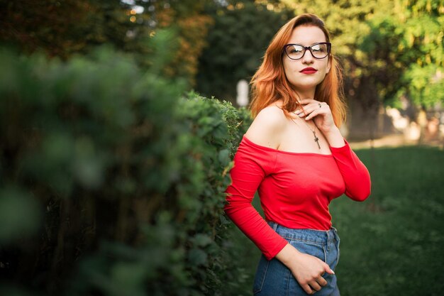 Привлекательная рыжеволосая женщина в очках, красной блузке и джинсовой юбке позирует в зеленом парке