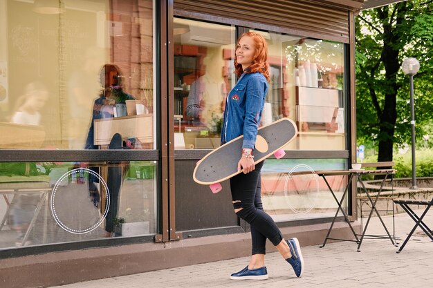 The attractive redhead female holds Longboard in the city park.