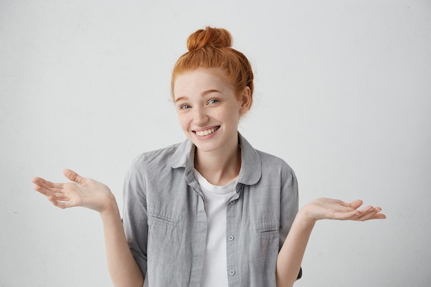 attractive redhead freckled teenager having broad smile shrugging her shoulders having some doubts. Emotional woman with red bun being confused while making some decisions