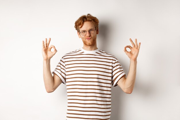 Attractive redhead guy in glasses showing okay signs and looking impressed, praising something cool, standing over white background