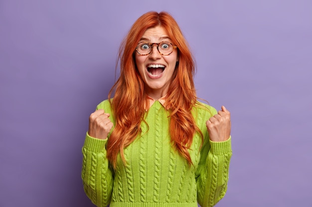 Foto gratuita attraente donna dai capelli rossi stringe i pugni con successo esclama e si sente trionfante essendo stupita non può credere nella sua vittoria indossa un maglione verde.
