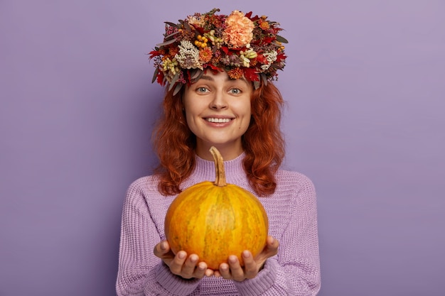 Attractive redhead woman wears autumnal wreath, holds ripe pumpkin, wears purple sweater.