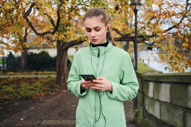 Attractive runner girl in earphones using cellphone resting in autumn city park