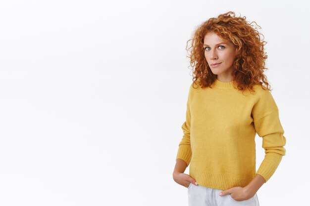 Привлекательная нахальная рыжая кудрявая женщина-предприниматель в желтом свитере, полуперевёрнутая стоит над белой стеной, руки в карманах джинсов, улыбается и смотрит в лицо уверенным, напористым взглядом