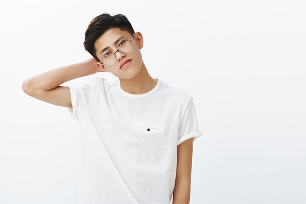 Attractive self-assured young asian with stylish hairstyle in glasses and white t-shirt tilting head touching back of neck making fashion pose gazing seriously