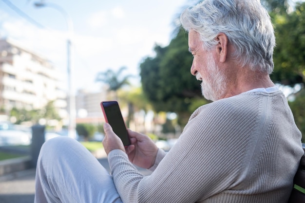 Привлекательный старший мужчина сидит на открытом воздухе в общественном парке с помощью мобильного телефона белые волосы улыбающиеся люди