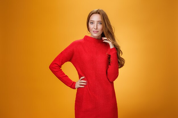 Attractive sensual and romantic redhead woman in red knitted elegant dress holding hand on hip smiling mysteriously as flirting playing with hair gazing at camera over orange background