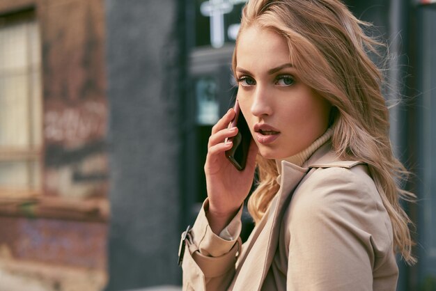 Attractive serious blond girl in trench coat talking on cellphone confidently looking in camera outdoor