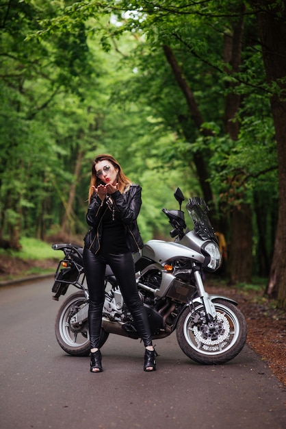 An attractive sexy girl dressed in leather posing near a sports motorbike outside