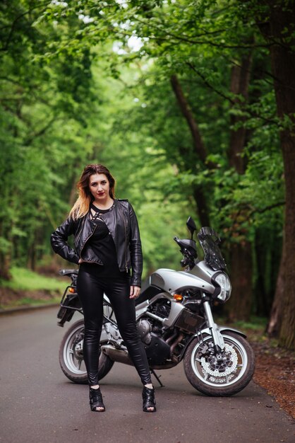 An attractive sexy girl dressed in leather posing near a sports motorbike outside