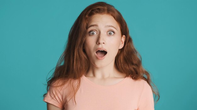 Attractive shocked redhead girl amazedly looking in camera with open mouth over colorful background Wow expression