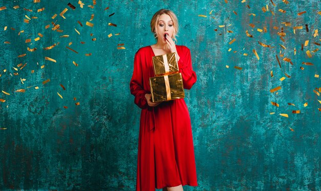Привлекательная потрясенная женщина в стильном красном платье празднует рождество и новый год с подарками