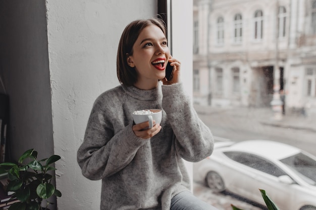 Привлекательная девушка с короткими волосами с красной помадой, одетая в серый свитер, разговаривает по телефону и держит чашку какао с зефиром у окна.