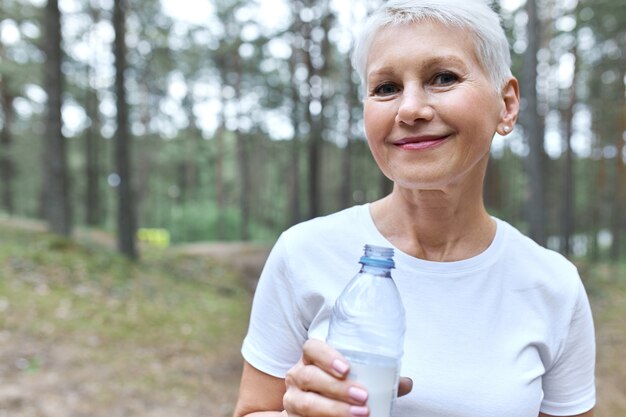 Attractive short haired middle aged woman in white t-shirt posing outdoors with pines