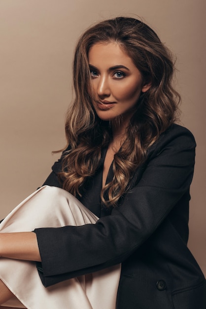 Attractive shy girl sitting alone and holding herself on the knees. Nude stylish makeup and curly long hairstyle. Black jacket and creamy dress on her body.