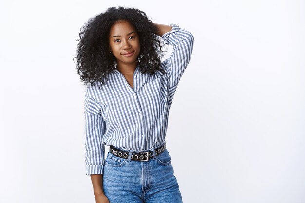 Attractive shy tender african american curly-haired female employee holding hand behind head unsure unconfident smiling friendly feeling awkward, uncomfortable new people around, white wall