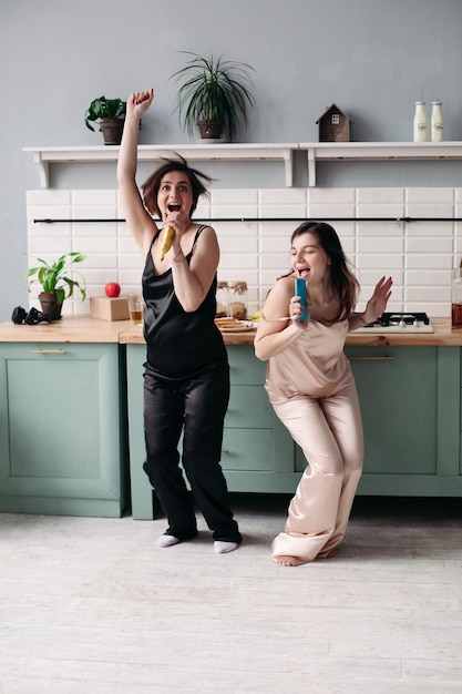 Free photo attractive sisters in black and white pajamas listening to music and dancing on kitchen in morning happy beautiful girls getting fun while cooking breakfast brunette friends singing and jumping