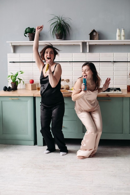 Attractive sisters in black and white pajamas listening to music and dancing on kitchen in morning Happy beautiful girls getting fun while cooking breakfast Brunette friends singing and jumping