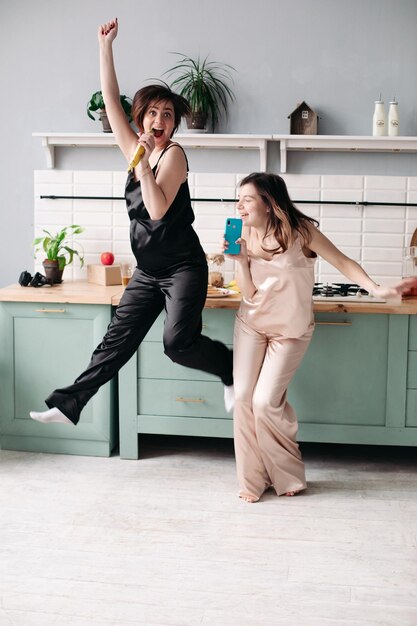 Attractive sisters in black and white pajamas listening to music and dancing on kitchen in morning Happy beautiful girls getting fun while cooking breakfast Brunette friends singing and jumping