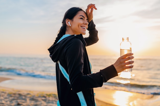 Привлекательная стройная женщина делает спортивные упражнения на утреннем пляже восхода солнца в спортивной одежде, пить питьевую воду в бутылке, здоровый образ жизни, слушает музыку на беспроводных наушниках, чувствуя себя горячим