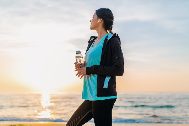 Attractive slim woman doing sport exercises on morning sunrise beach in sports wear, thirsty drinking water in bottle, healthy lifestyle, listening to music on wireless earphones, smiling happy