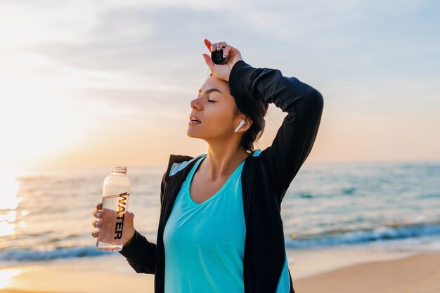 Привлекательная стройная женщина делает спортивные упражнения на утреннем пляже восхода солнца в спортивной одежде, пить питьевую воду в бутылке, здоровый образ жизни, слушает музыку на беспроводных наушниках, улыбается счастливым