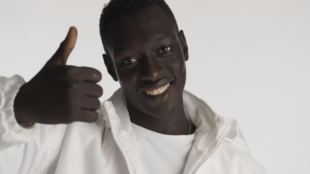 カジュアルな服装で魅力的な笑顔のアフリカ系アメリカ人の男性は、白い背景の上に幸せそうに見える親指を維持します。好きな表現