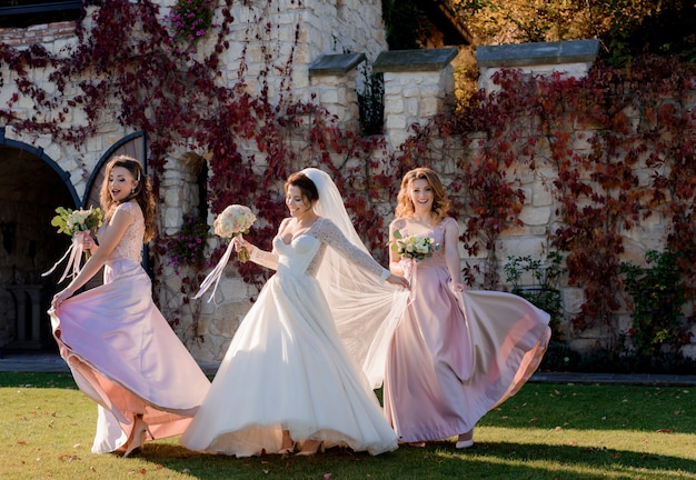 Attractive smiling bride and bridesmaids are dancing and having fun in front of stone building covered with red ivy