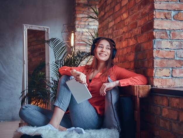 An attractive smiling brunette girl dressed in casual clothes using a tablet while listening to exciting music via good headphones. Relaxing in a room with loft interior.