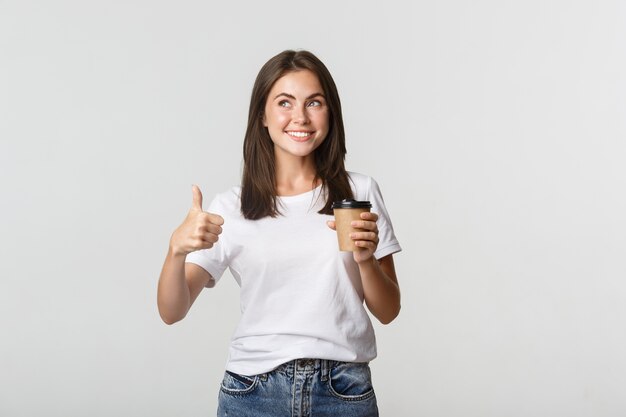 Привлекательная улыбающаяся брюнетка девушка выглядит довольной, пьет кофе и показывает палец вверх, рекомендую кафе.