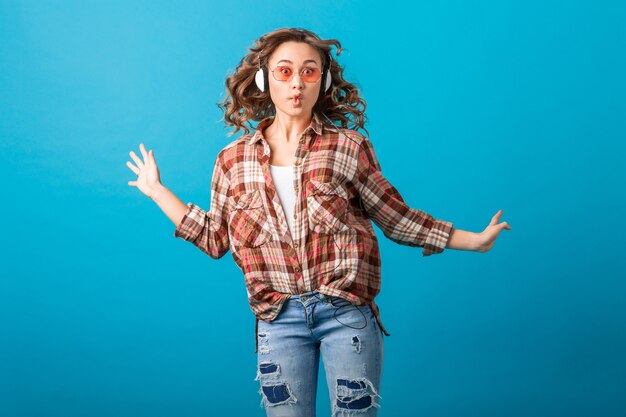 Attractive smiling emotional woman jumping with funny crazy face expression in checkered shirt and jeans isolated on blue studio background, wearing pink sunglasses