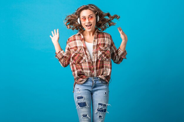 Attractive smiling emotional woman jumping with funny crazy face expression in checkered shirt and jeans isolated on blue studio background, wearing pink sunglasses