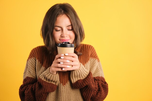 黄色の背景の上に夢のようなコーヒーの香りがする居心地の良いセーターの魅力的な笑顔の女の子